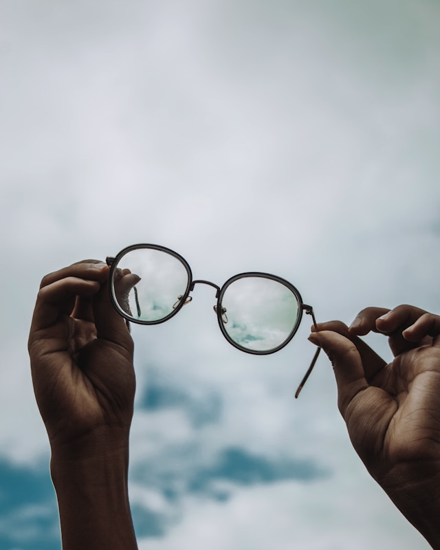 A pair of round eyeglasses held up against a bright cloudy sky